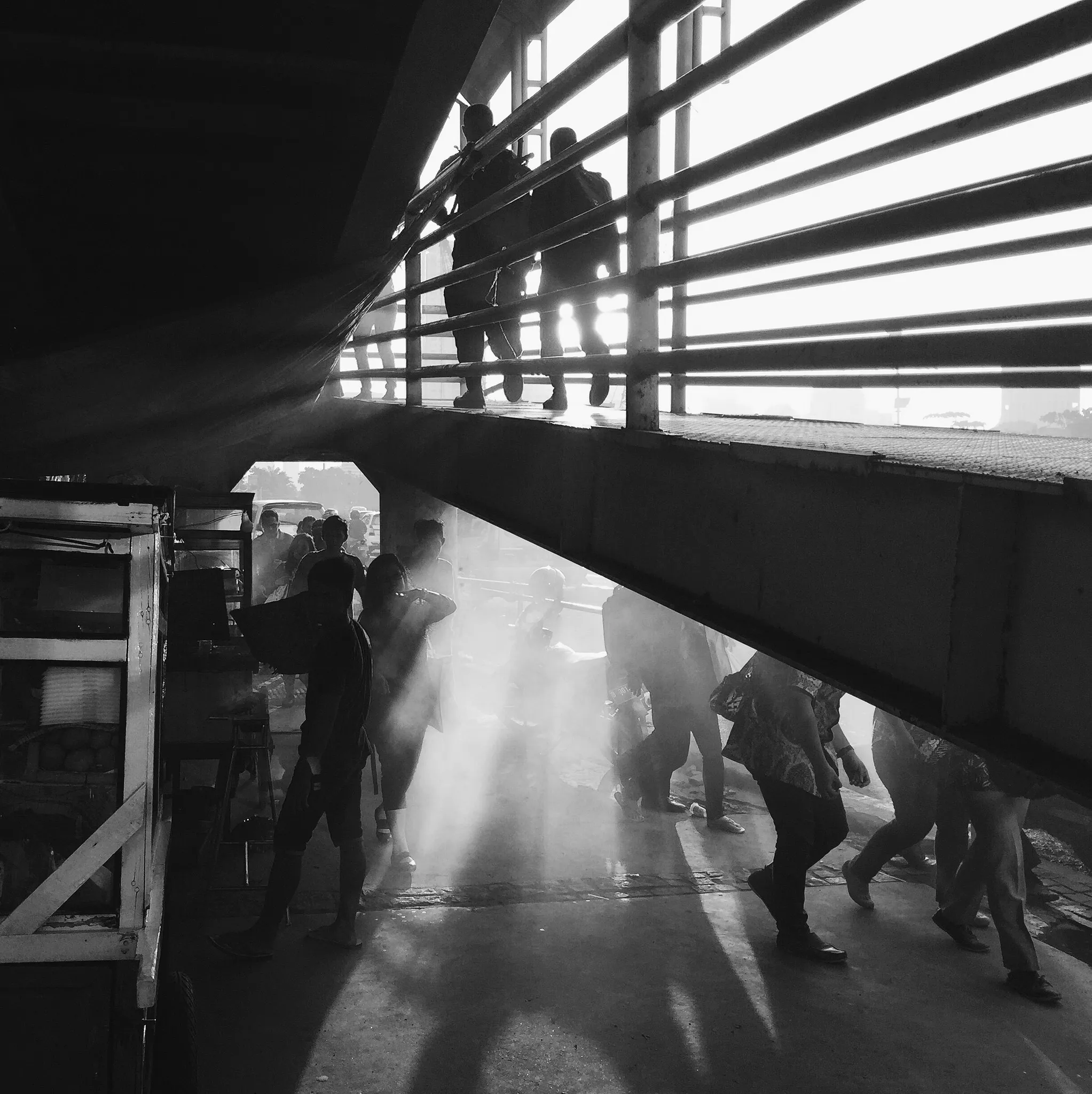 People walking through shafts of light beneath a bridge, in dramatic black and white.