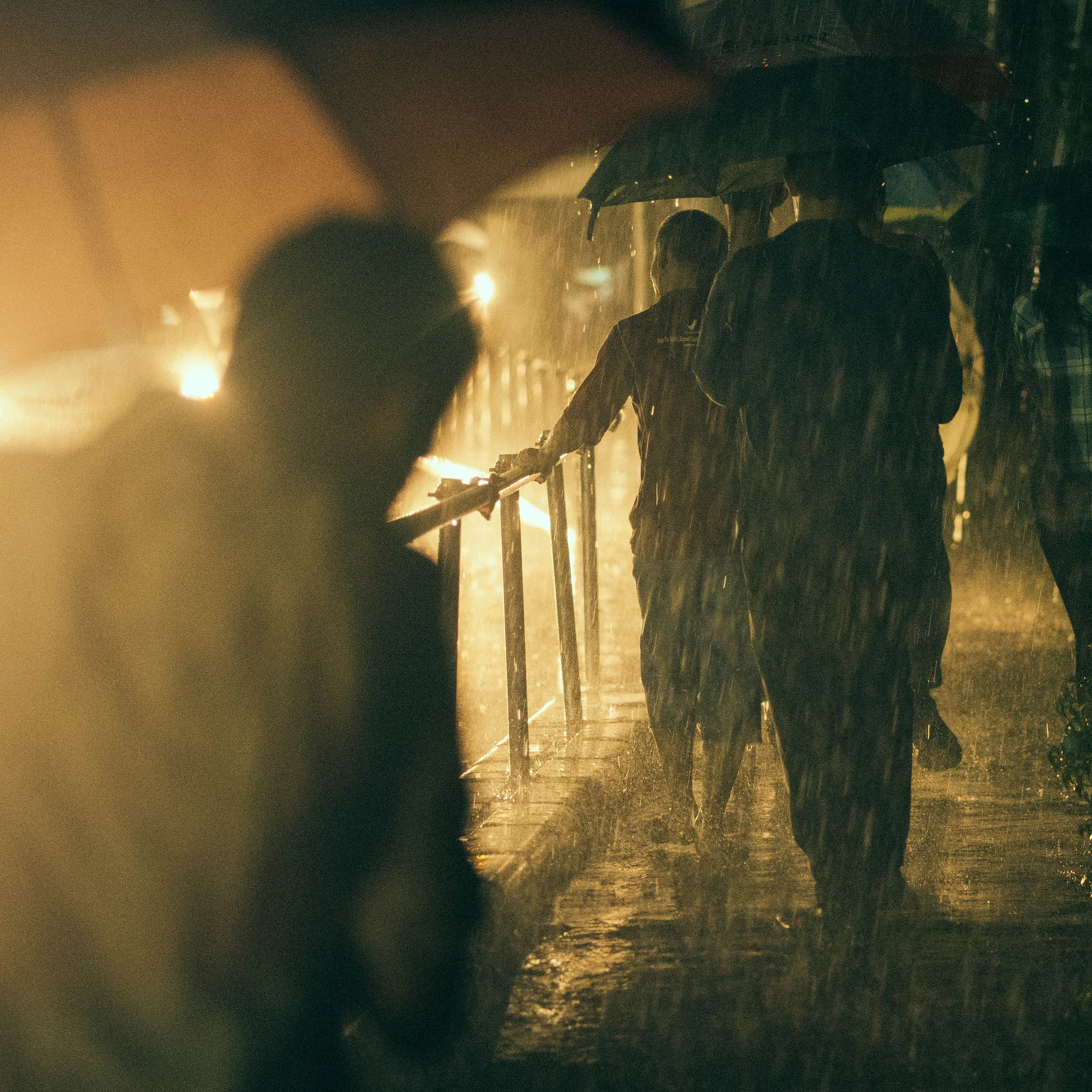 Heavy rain at night, people with umbrellas walk past a railing.