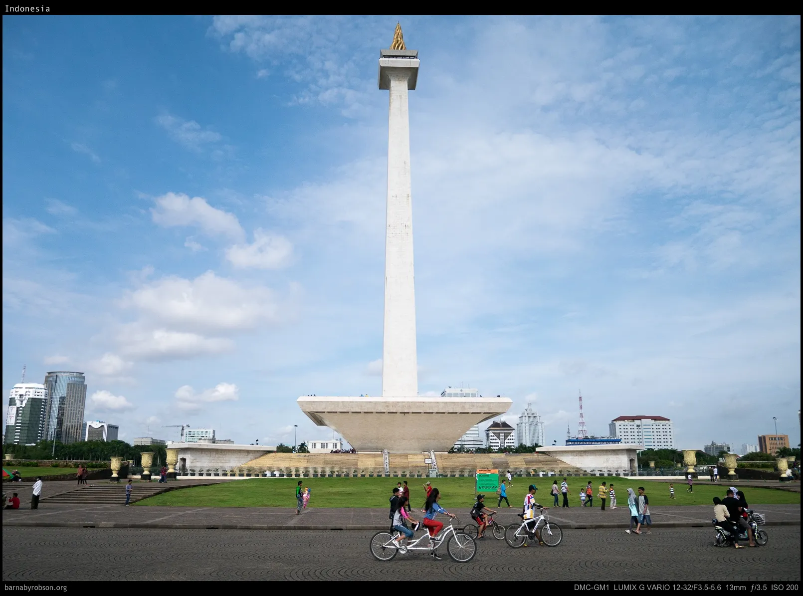 White monument with golden flame, people cycling and walking in park.