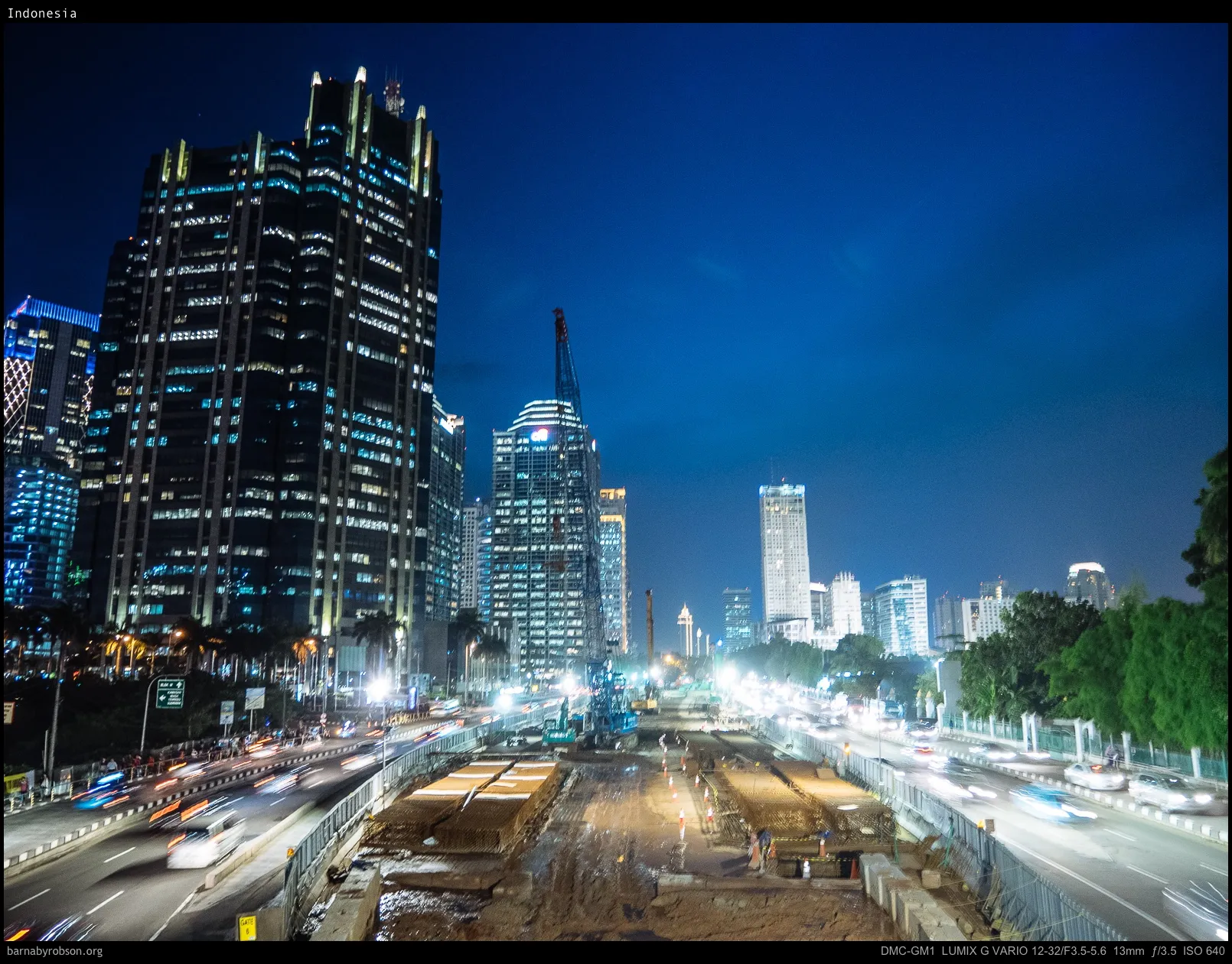 Night view of city skyscrapers, busy road, and construction site with crane.
