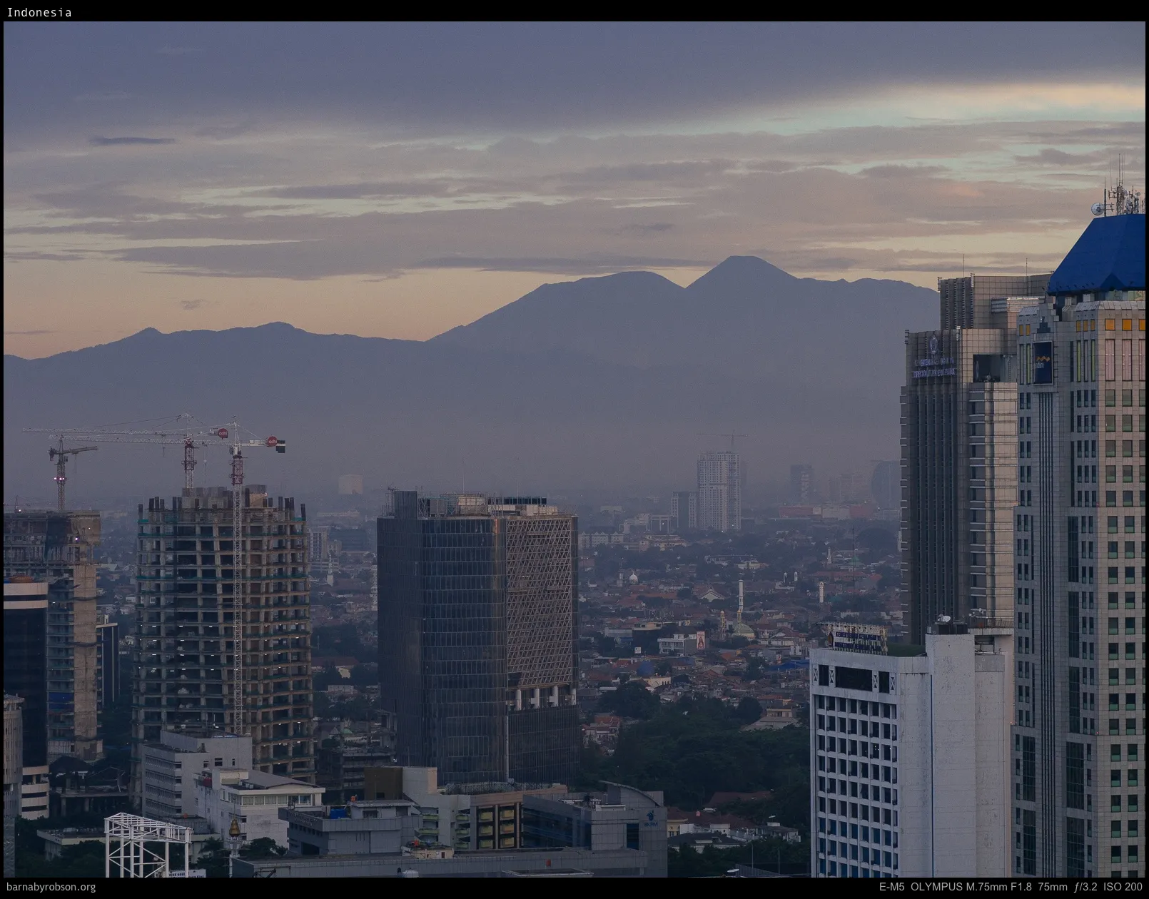 Hazy city skyline with tall buildings, construction cranes, and distant mountains at dusk.