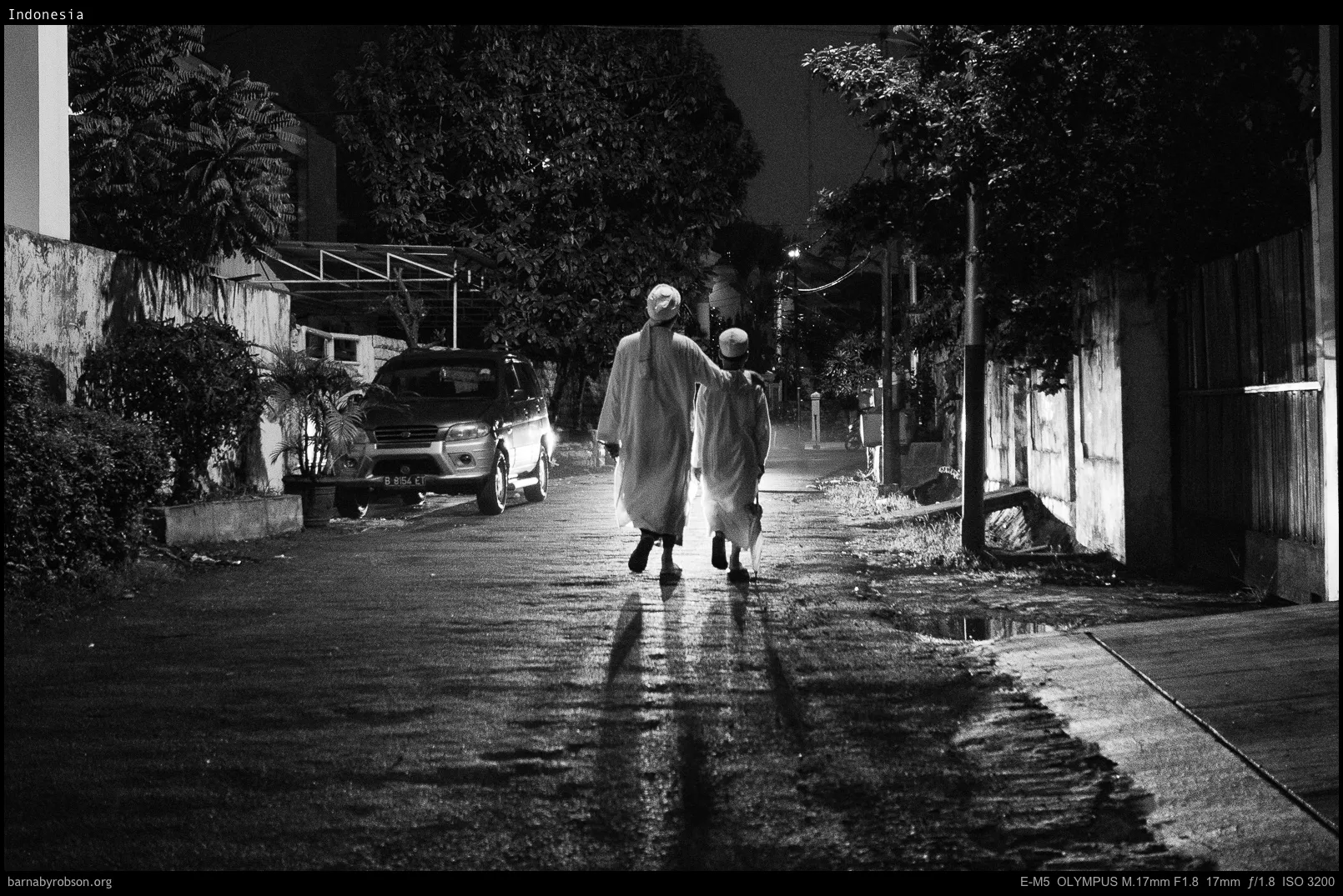 Black and white: two robed figures walk down a wet street at night.