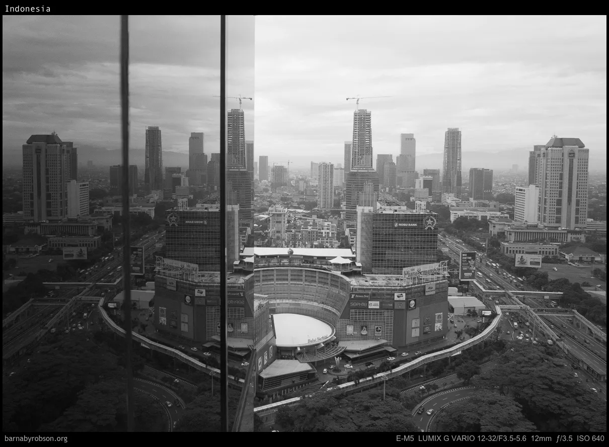 Black and white cityscape: skyscrapers, traffic, Plaza Semanggi mall, and window reflection.