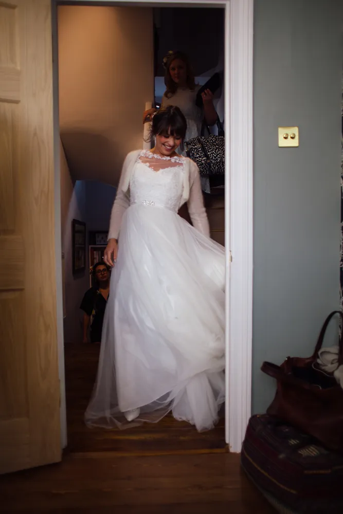 Smiling bride in white dress and cardigan walking through a doorway.