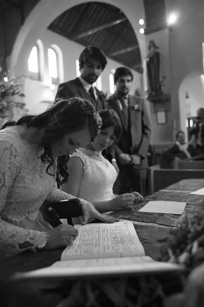Two brides signing a wedding register, with two men in suits watching.