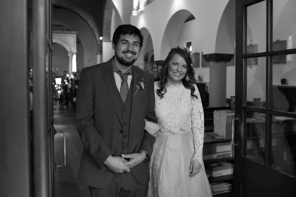Smiling bride in lace and groom in suit, arm-in-arm, in church.