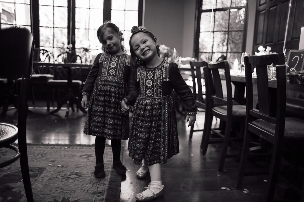 Two smiling girls in patterned dresses holding hands indoors.
