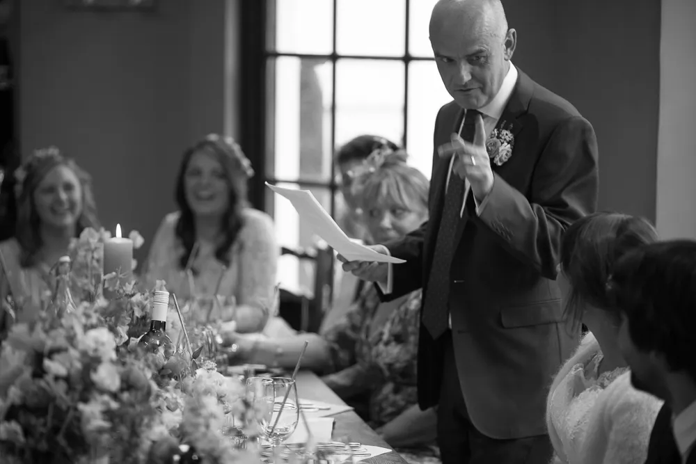 Man in suit speaks to smiling guests at a floral-decorated table.