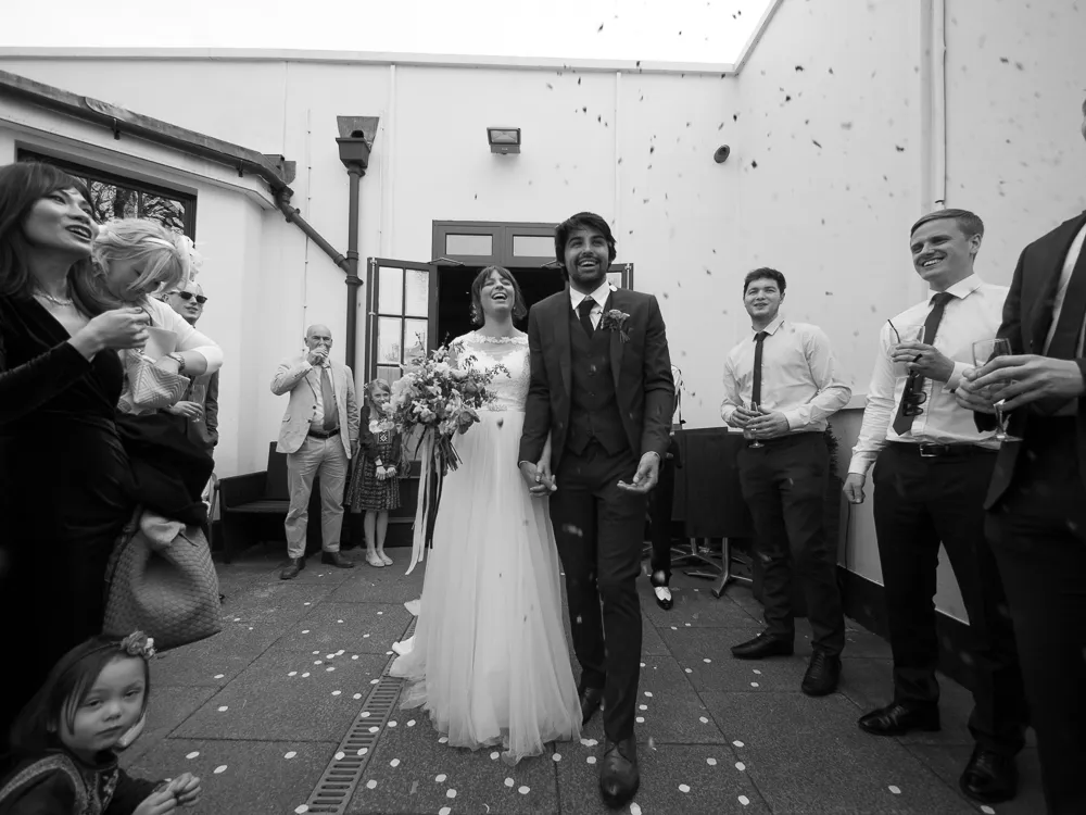Smiling bride and groom walk through confetti, surrounded by wedding guests.