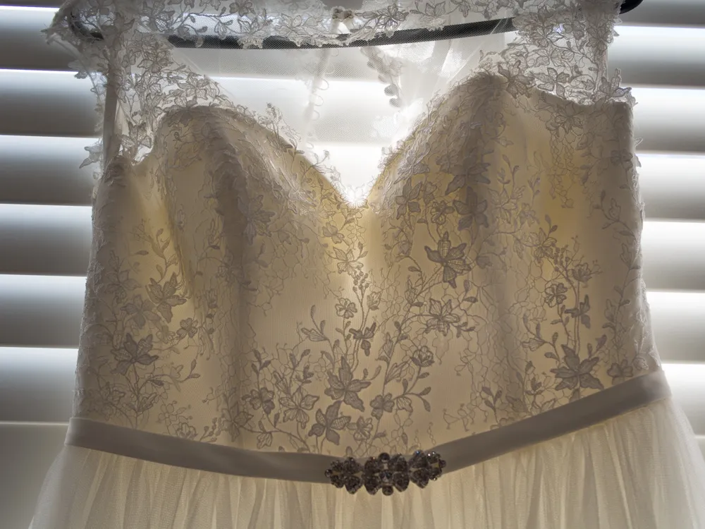 Ivory lace wedding dress with a sparkling belt, backlit by blinds.