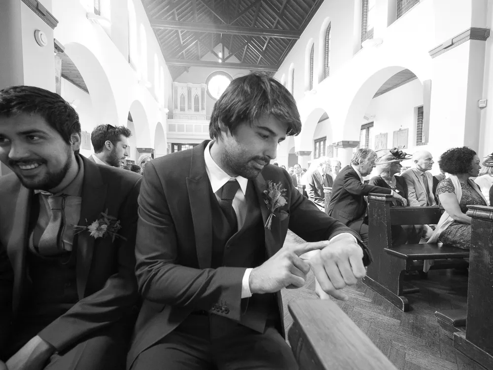 Two men in suits in a church pew; one smiles, one checks his watch.