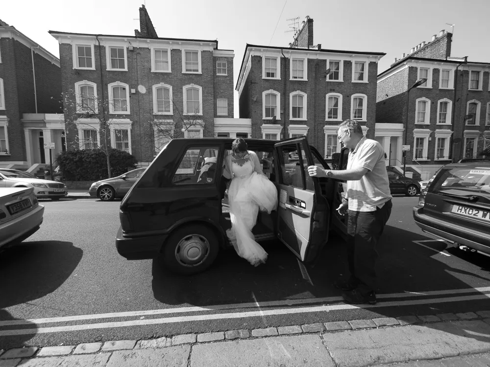 Bride in white dress exiting car, man holding door, street, brick buildings.