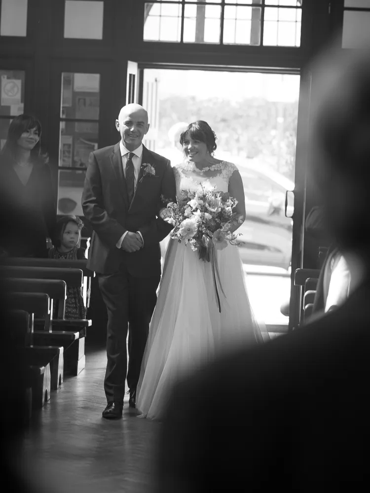 Smiling bride holding bouquet walks down aisle with escort, black and white.