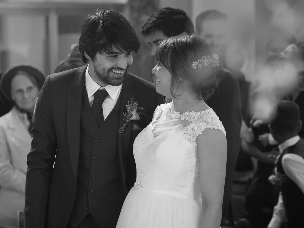 Smiling bride in lace dress and groom in suit, looking at each other.