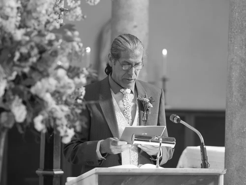Man in suit and glasses reading a document at a lectern.