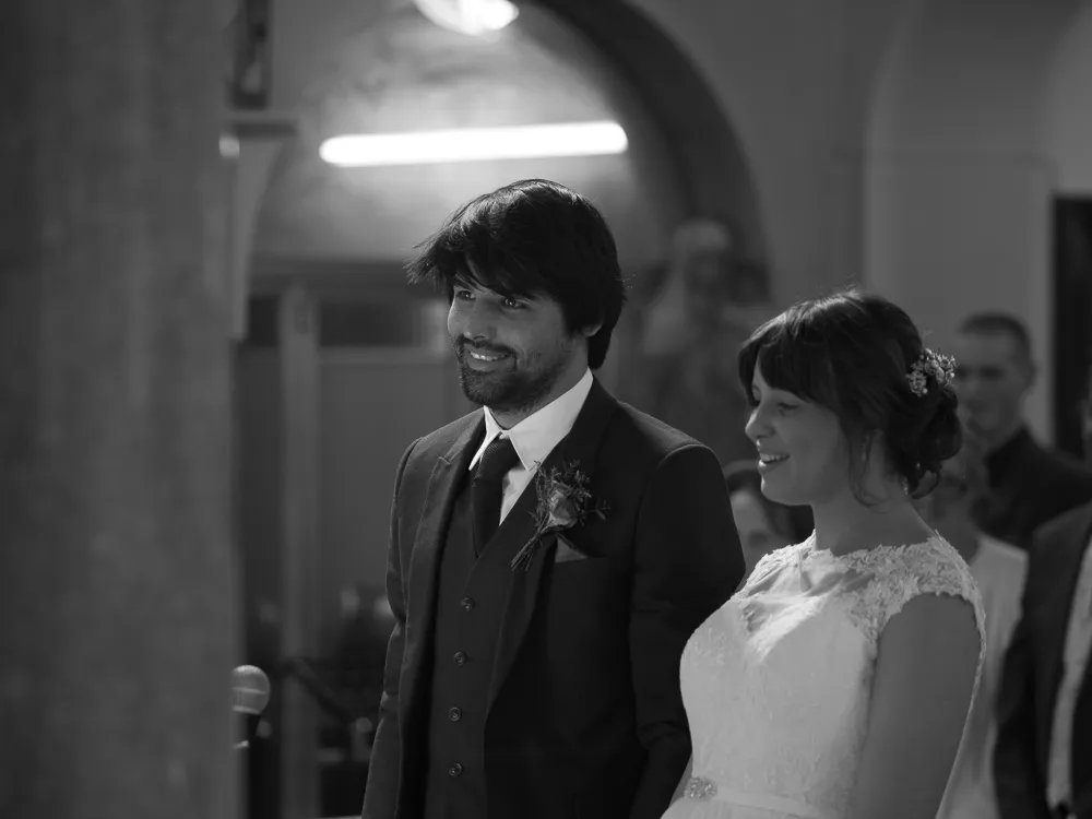 Smiling bride in white lace dress and groom in dark suit, black and white.