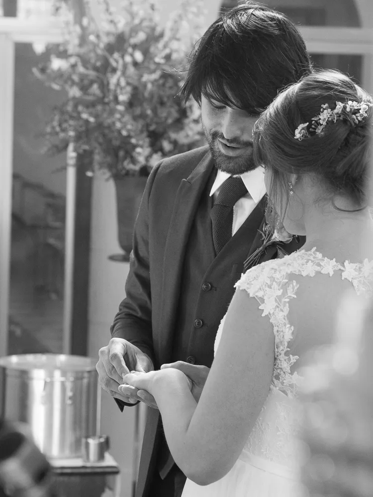 Groom places wedding ring on bride's finger in black and white.