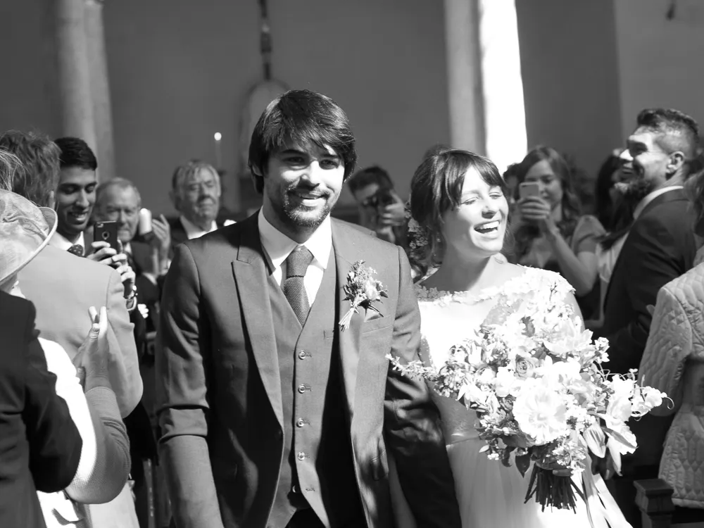 Smiling bride and groom walk past guests, she holds a large bouquet.
