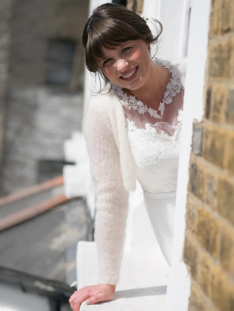 Smiling bride in white dress and cream cardigan leaning from window.