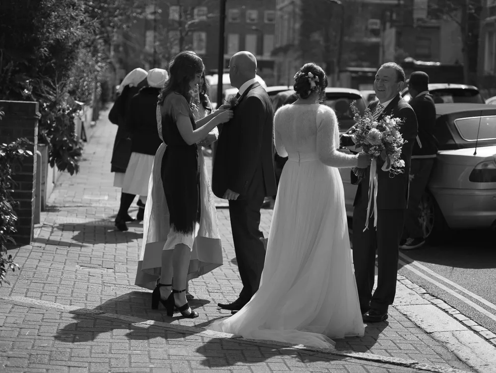 Black and white: woman pinning boutonnière, bride with bouquet on pavement.
