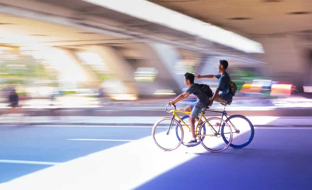 Two young men cycling under a bridge, one pushing the other.