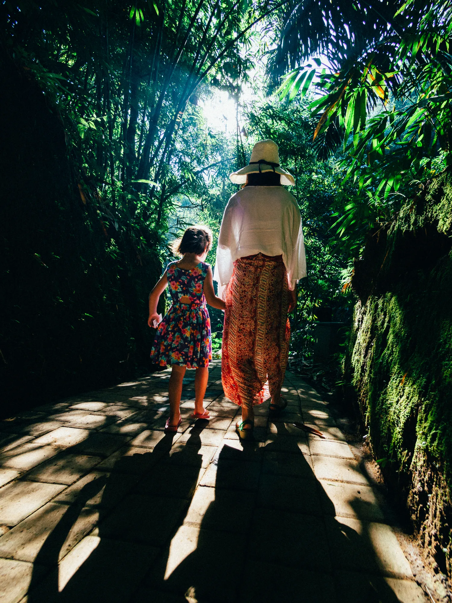 Woman and child walk on a paved path, sun streaming through dense green trees.