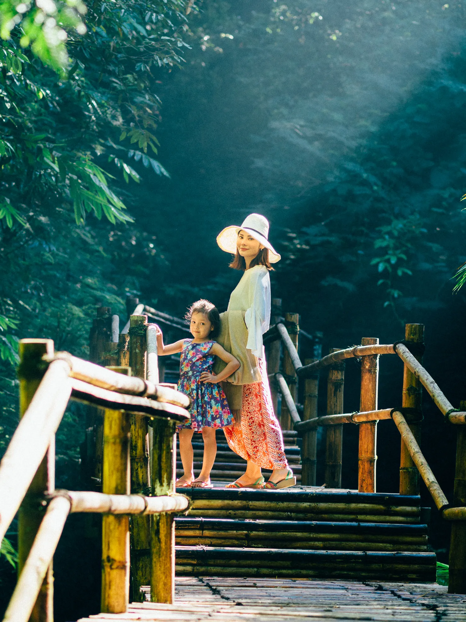 Woman and girl on bamboo bridge, sunbeams filtering through trees.