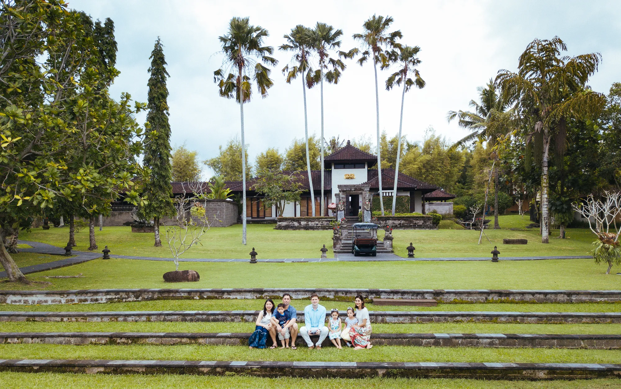 Family on stone steps in a tropical garden with a traditional Balinese building.