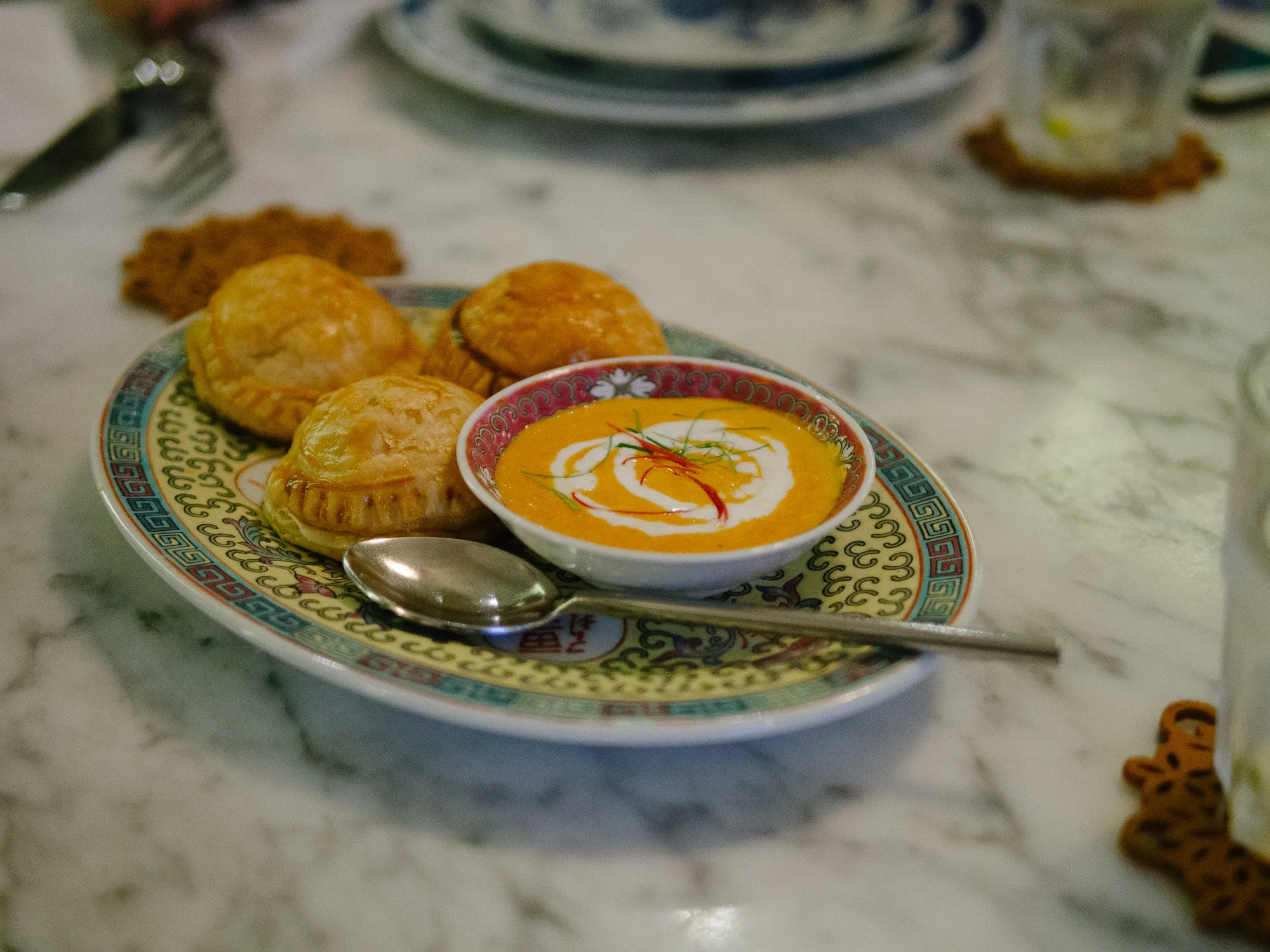 Four golden pastries and orange soup with garnish on a decorative plate.