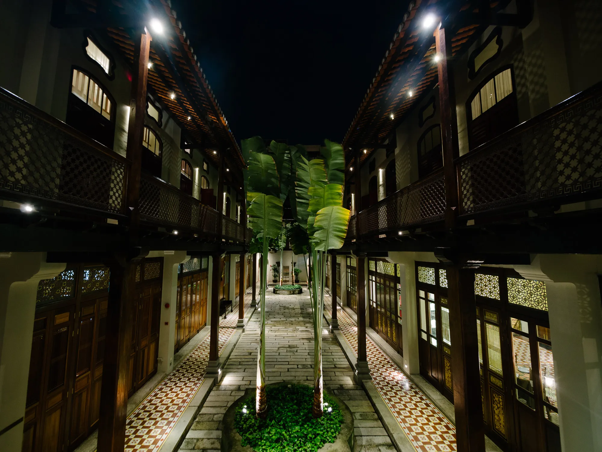 Symmetrical traditional courtyard at night, illuminated, with balconies, pathway, and plants.