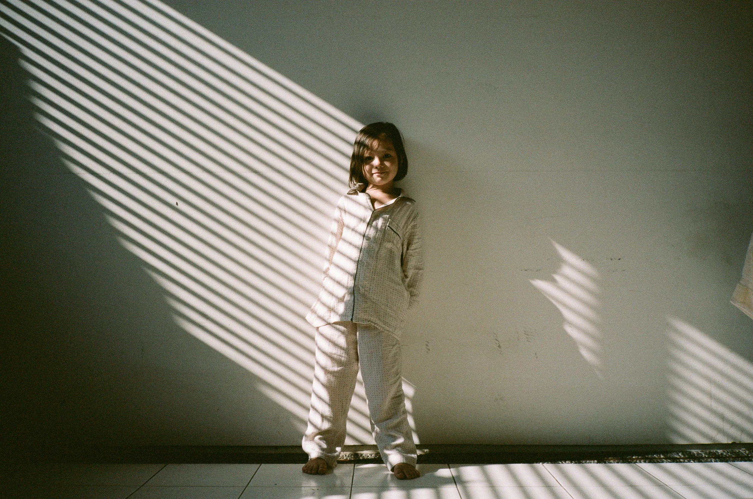 Barefoot child in pyjamas stands against a wall with striped sunlight shadows.