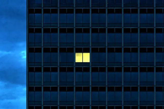 A dark office facade at dusk with a single lit window — one bright room in the grid.
