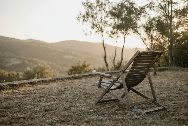A weathered wooden deckchair sitting alone on a quiet grassy terrace overlooking hazy hills at golden hour — work less, enjoy more.