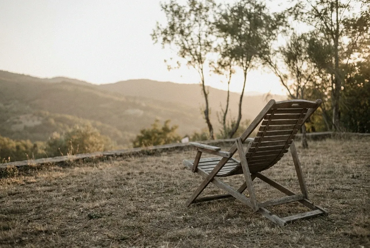 A weathered wooden deckchair sitting alone on a quiet grassy terrace overlooking hazy hills at golden hour — work less, enjoy more.