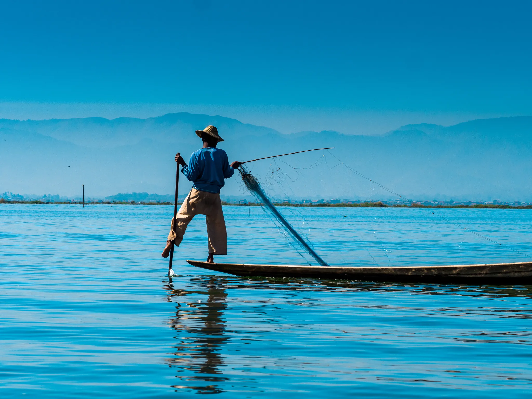 Fisherman leg-rowing a boat, casting a net on a blue lake.
