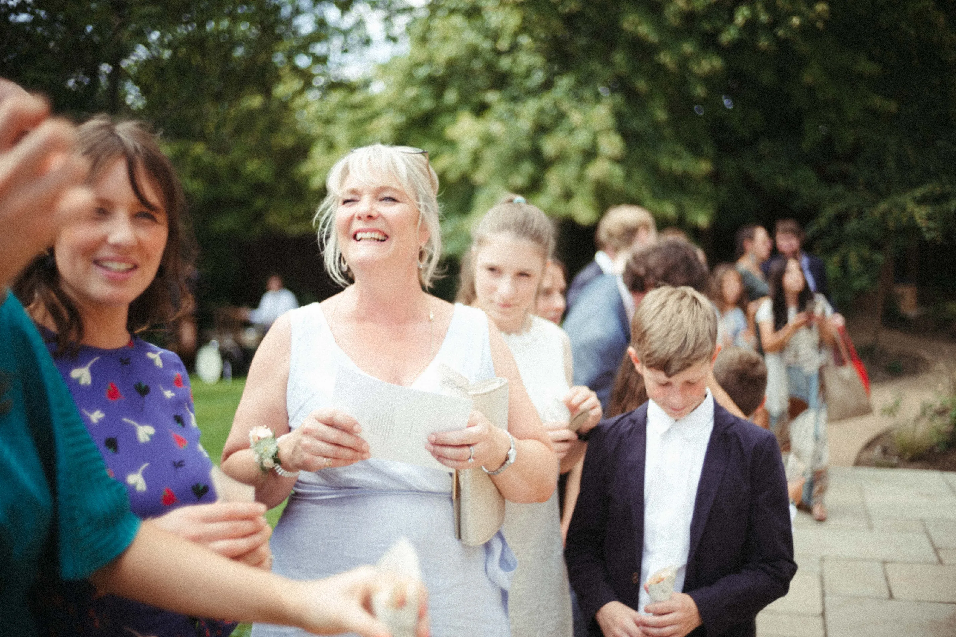 Smiling woman in light blue dress holding paper, with guests outdoors.