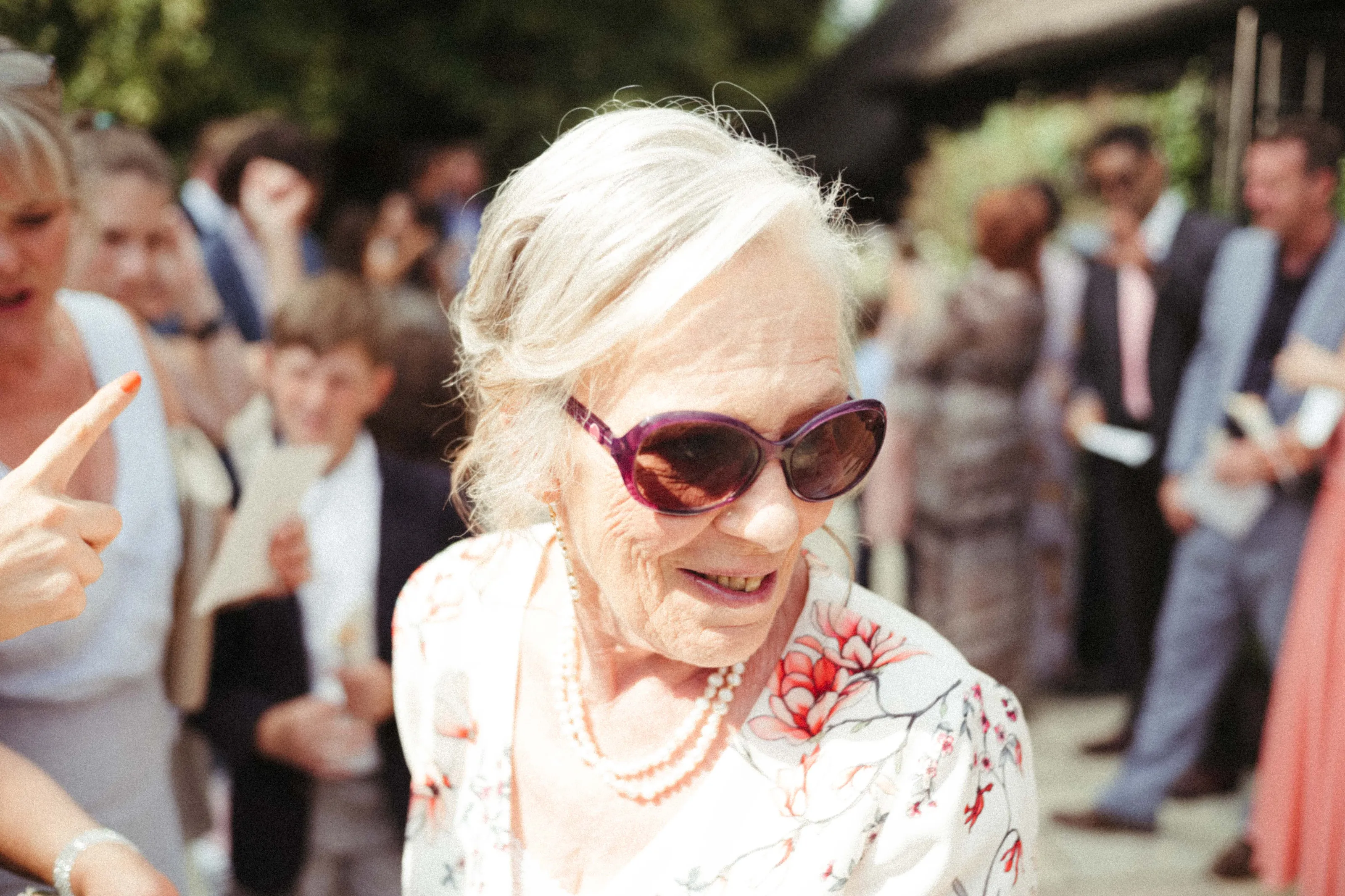Smiling older woman in purple sunglasses, pearls, and floral top at outdoor gathering.