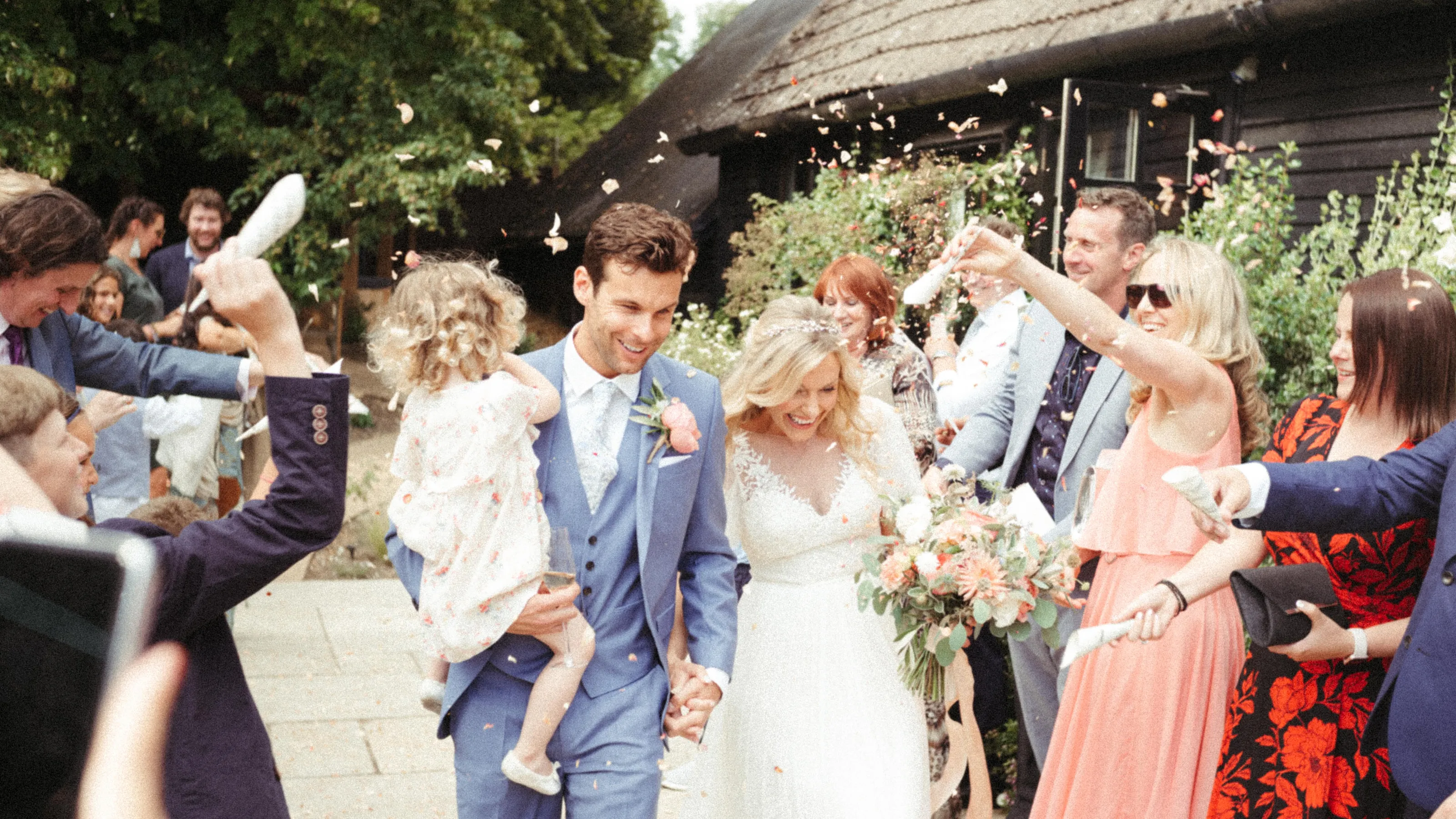Smiling bride and groom, groom holding child, showered with confetti.