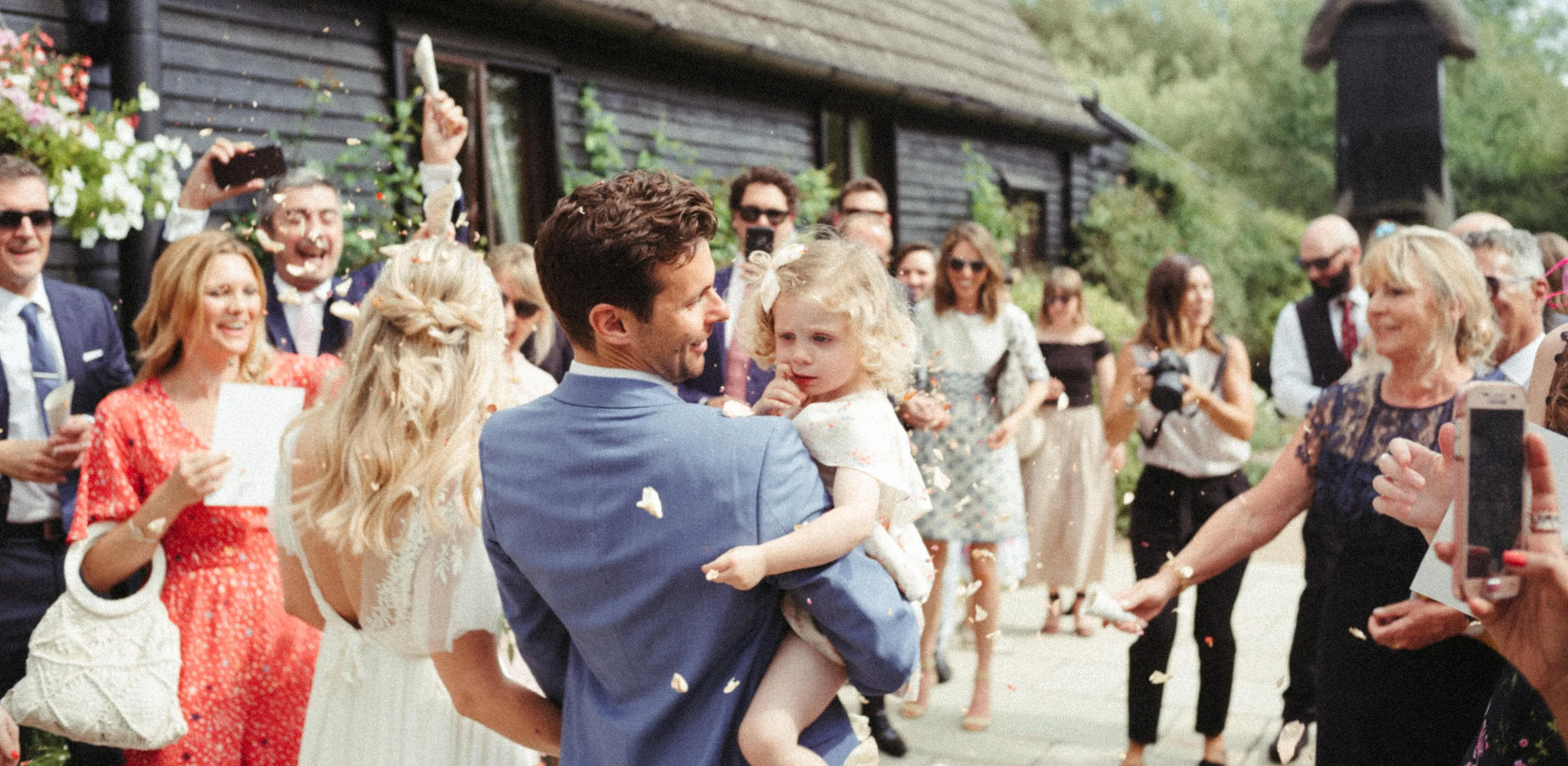 Wedding guests throw confetti over groom holding child and bride.