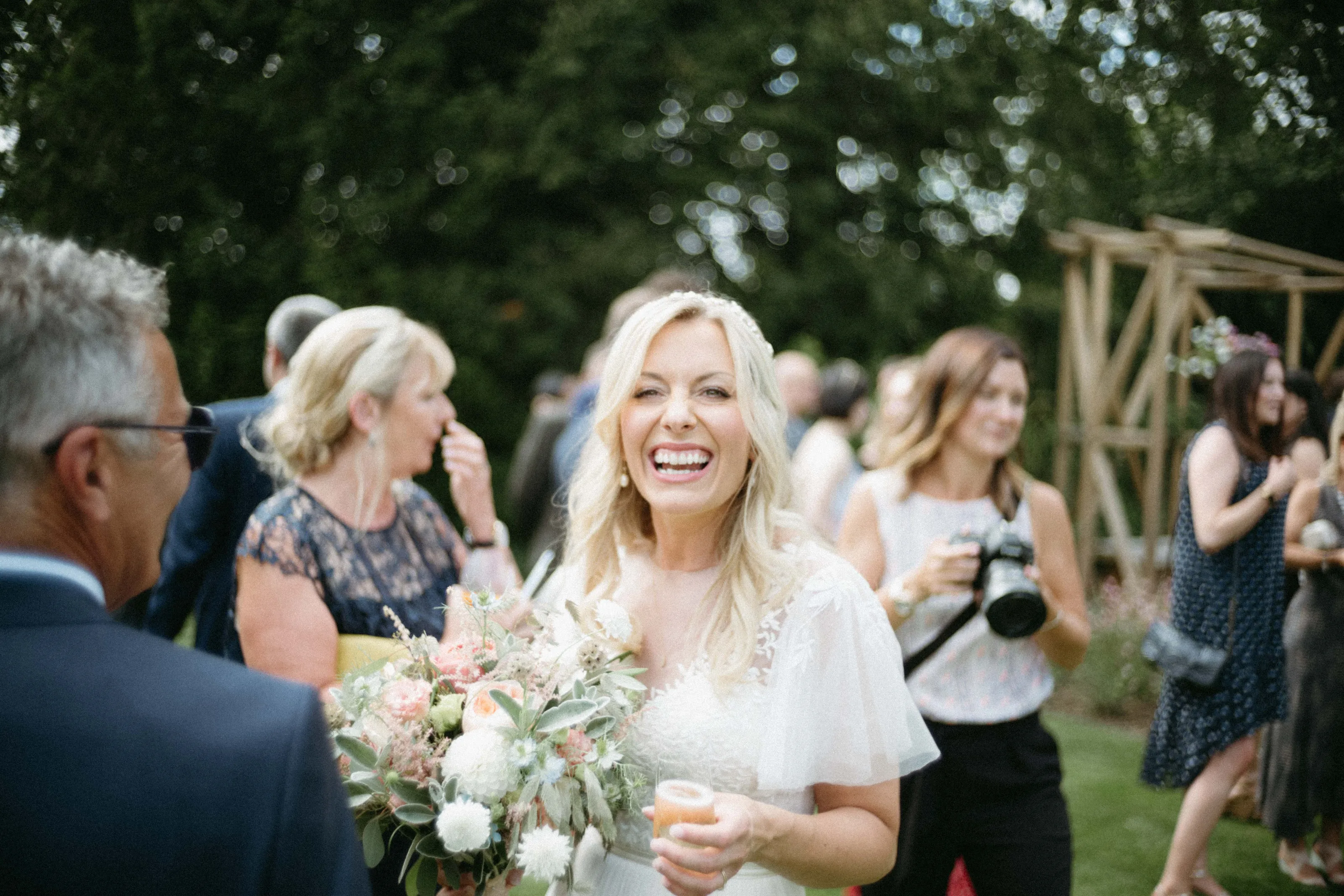 Laughing bride holding bouquet and drink at outdoor wedding with guests.