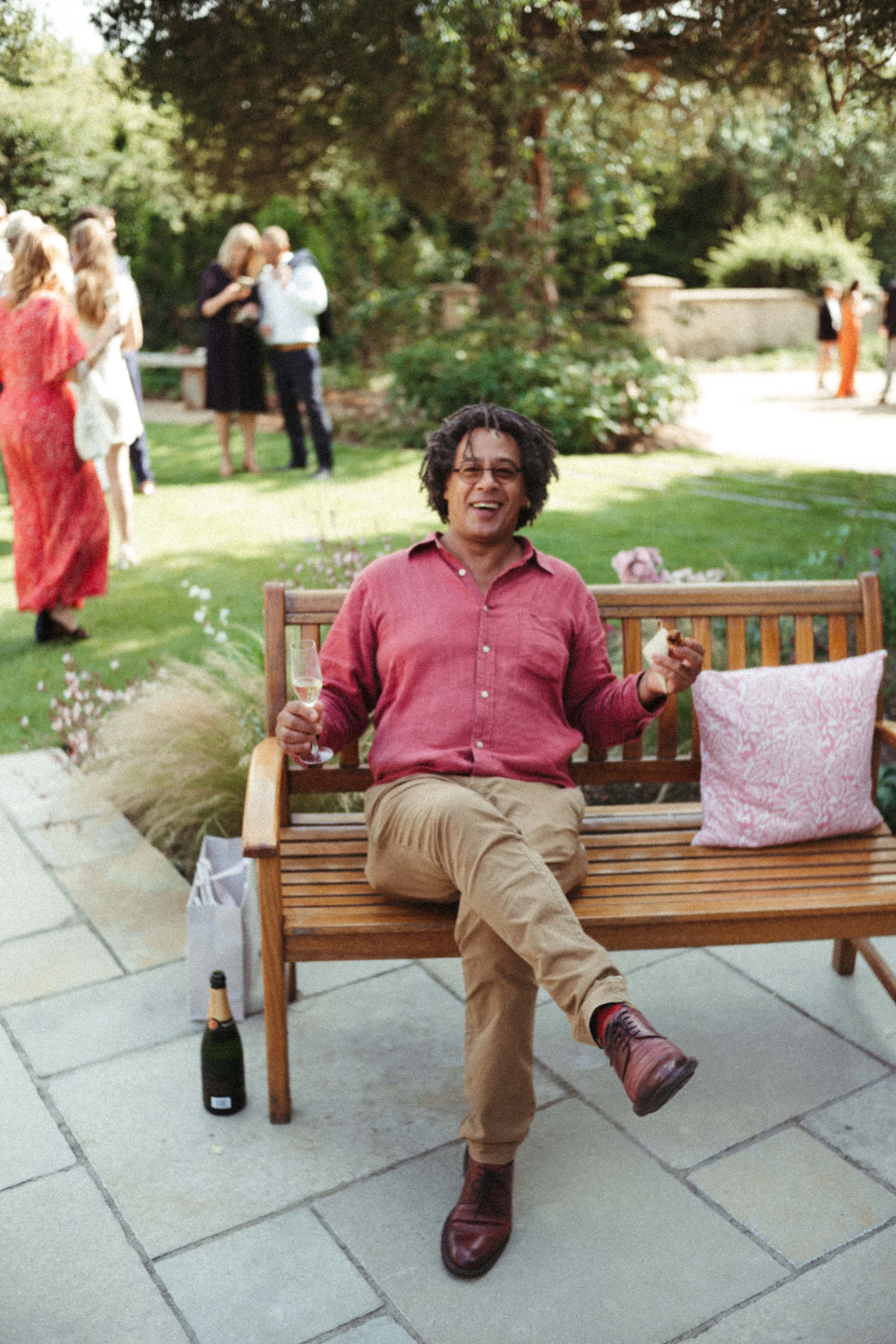 Smiling man on garden bench with champagne and food, guests in background.