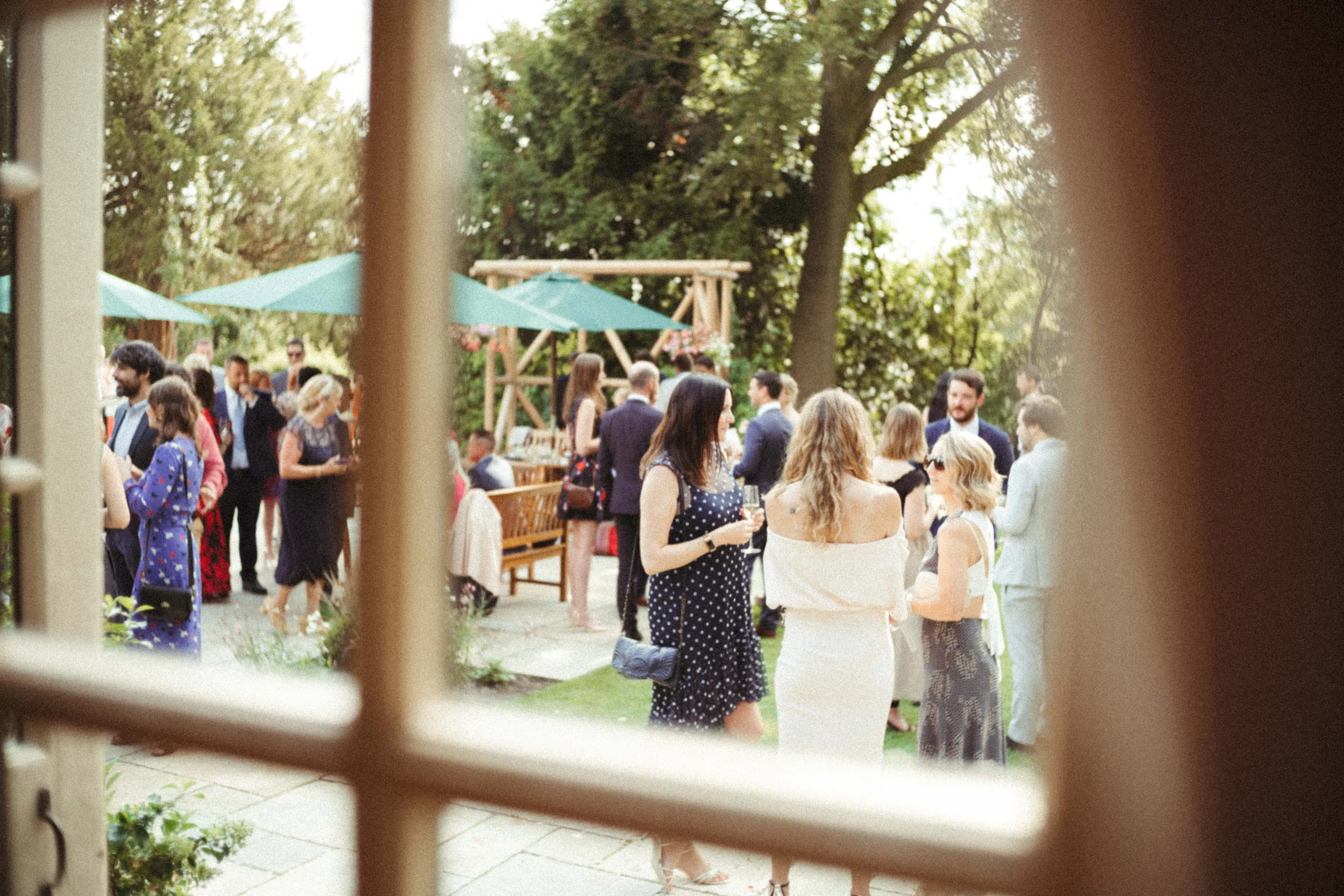 Guests socialising outdoors in a garden with green umbrellas, framed by a window.