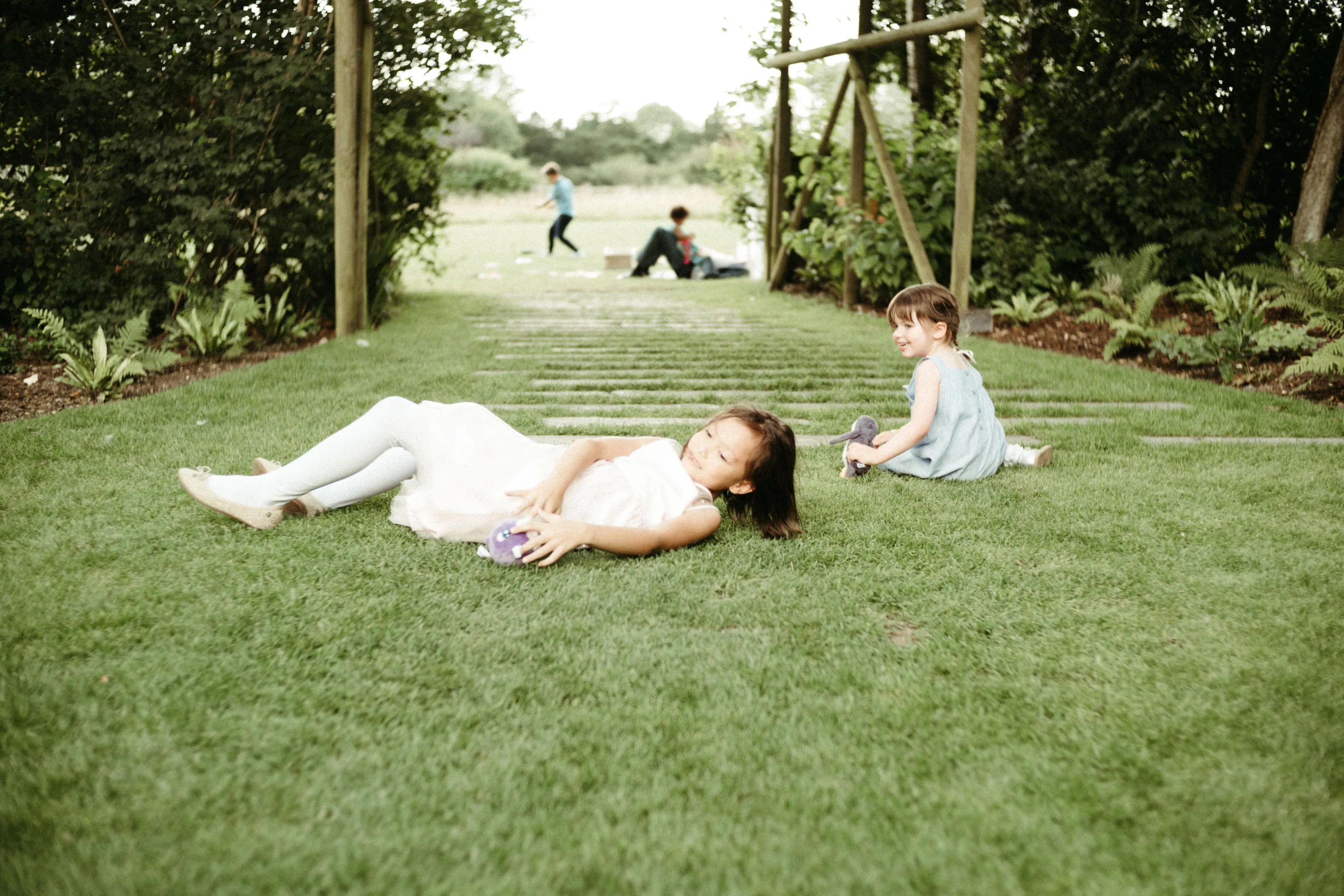 Two girls lie and sit on grass, playing with toys.