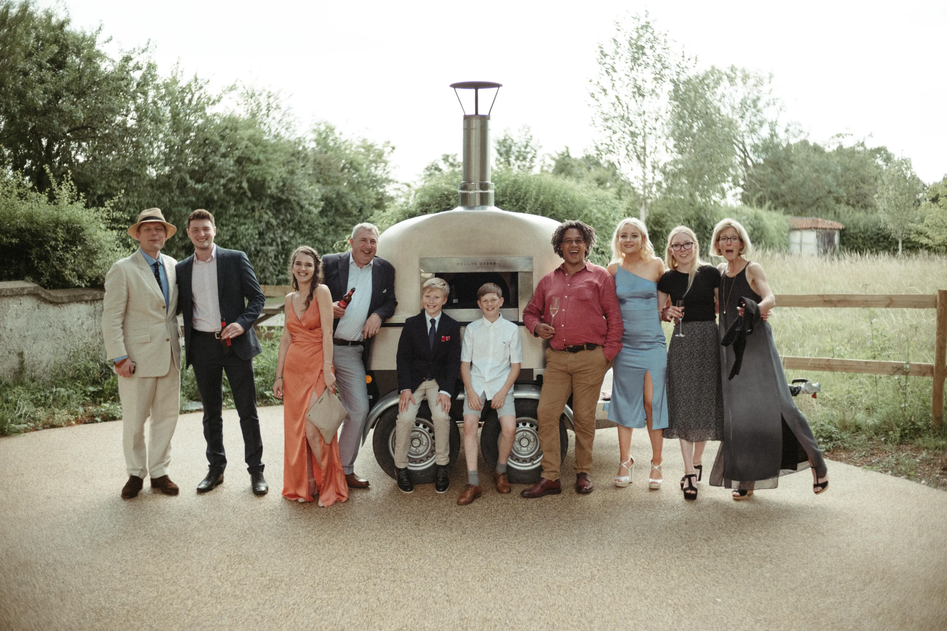 Eleven smiling people gathered around a mobile pizza oven outdoors.