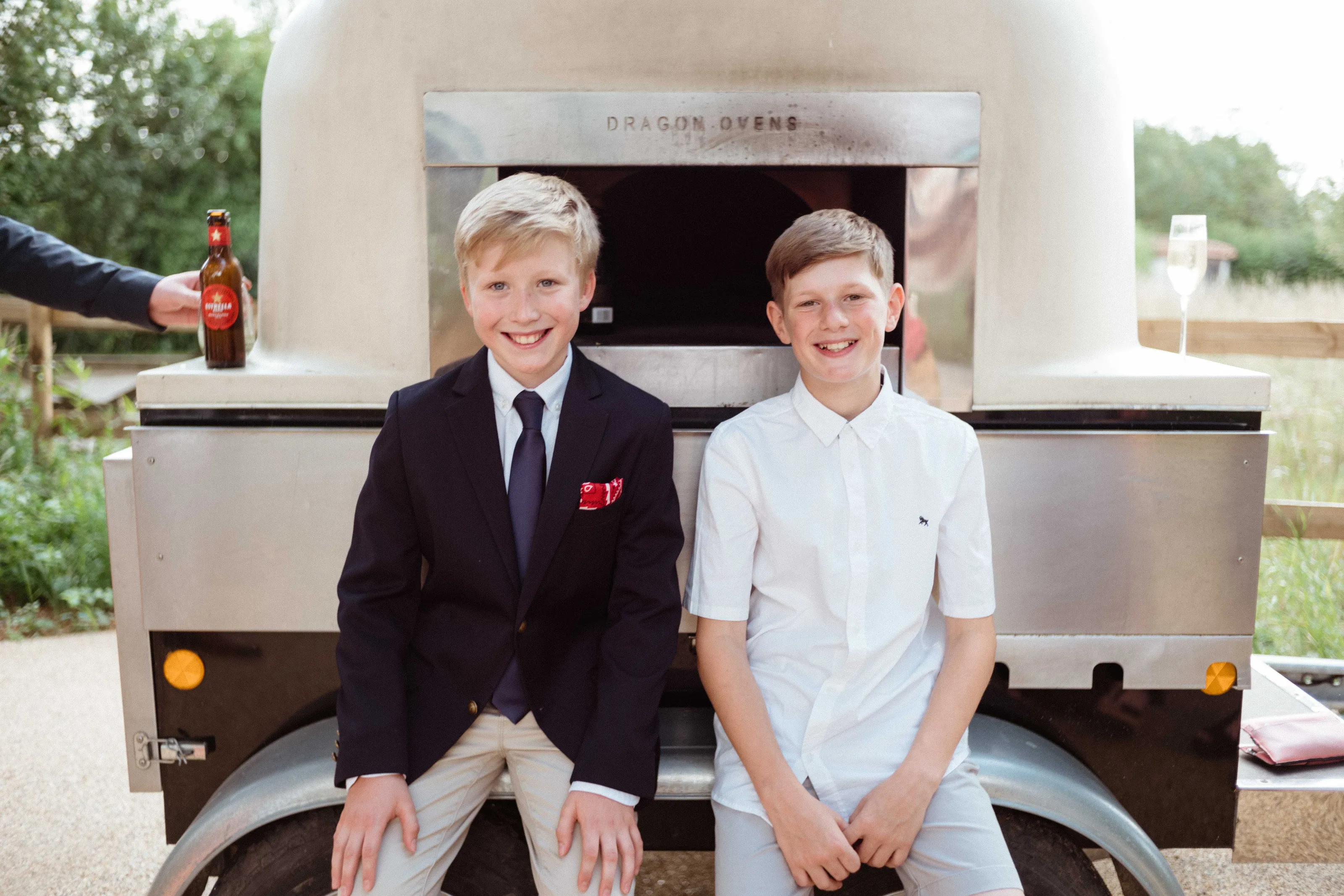 Two smiling boys sit in front of a 'Dragon Ovens' pizza oven trailer.