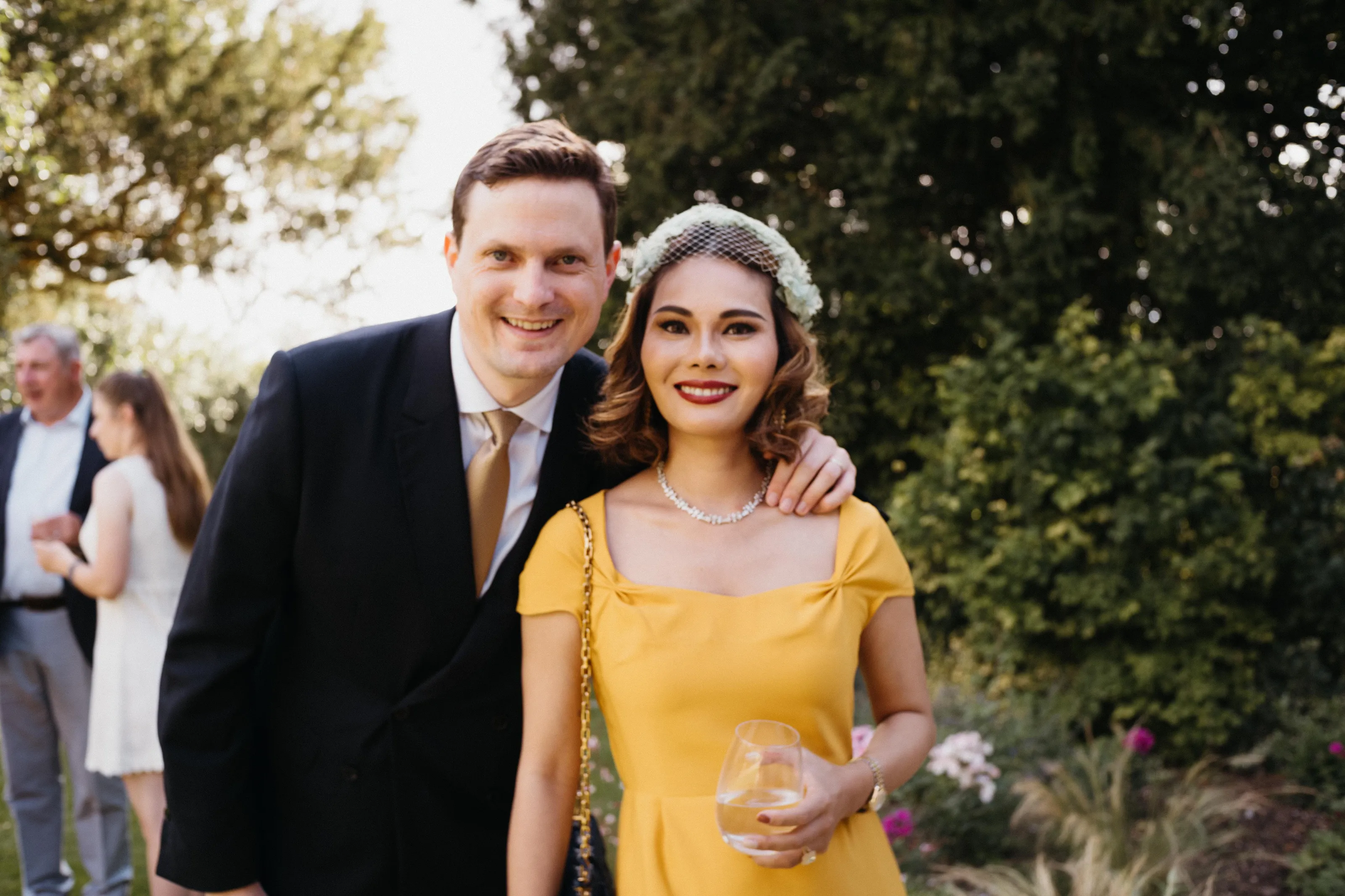 Smiling man in suit and woman in yellow dress with wine outdoors.
