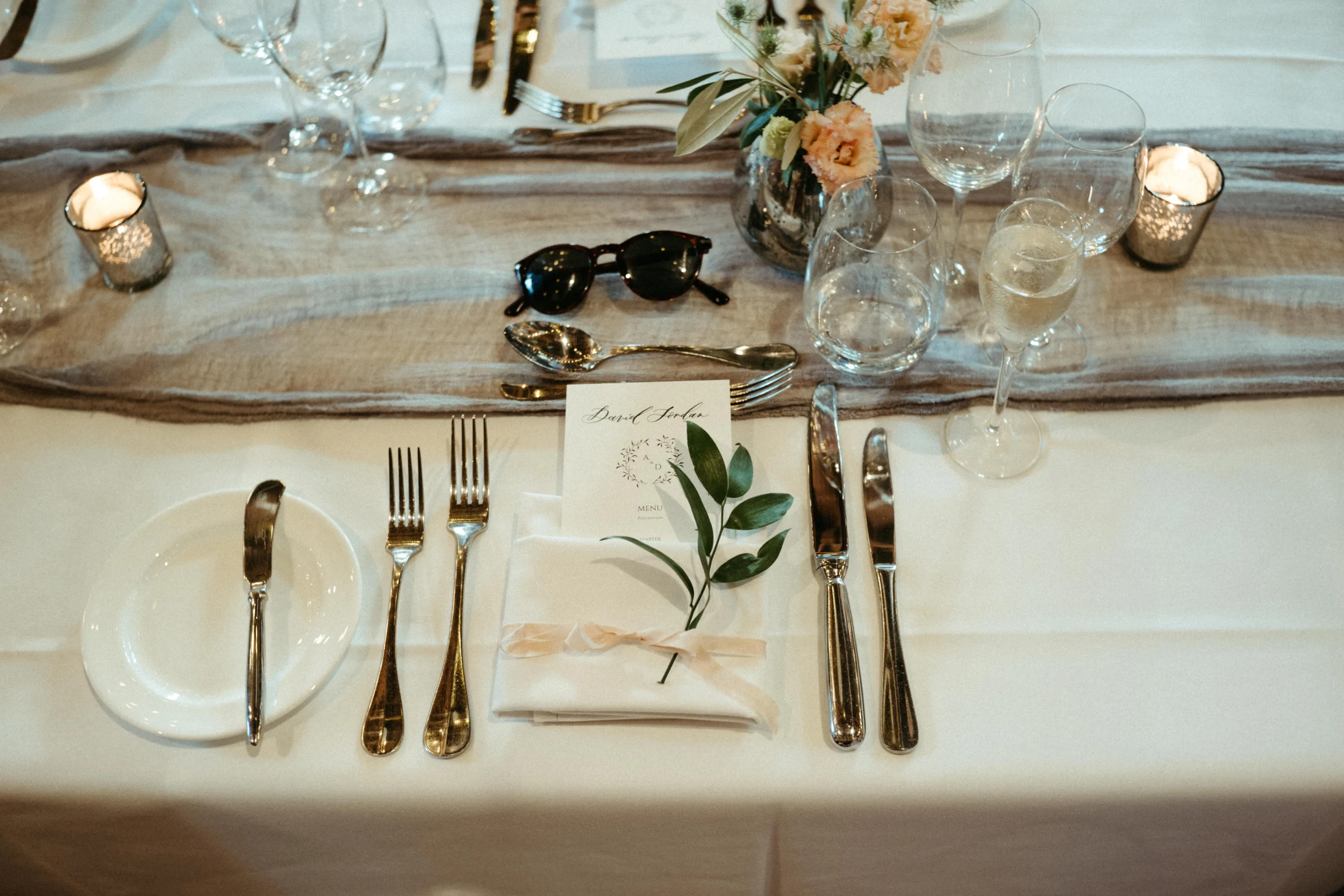Table setting with gold cutlery, menu, flowers, candles, glasses, and sunglasses.