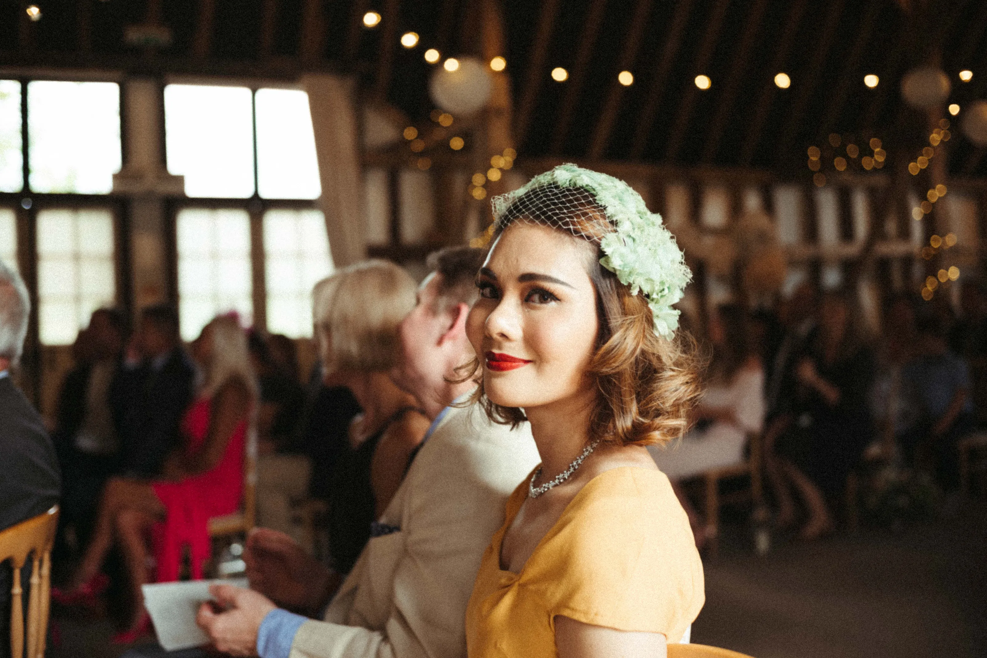 Woman in yellow dress, green fascinator, red lipstick, at a festive event.