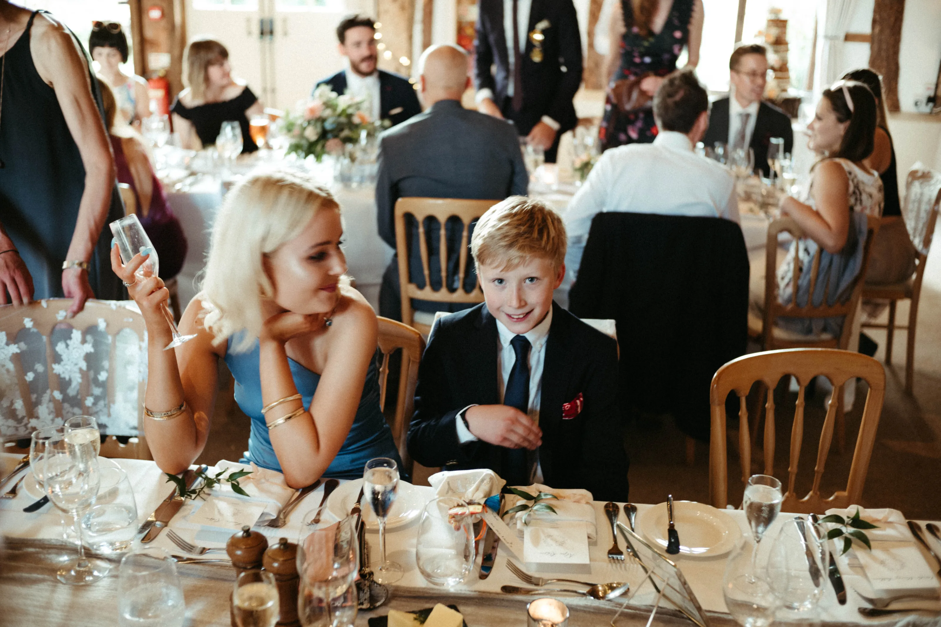 Smiling blonde boy in suit, woman in blue dress at reception table.