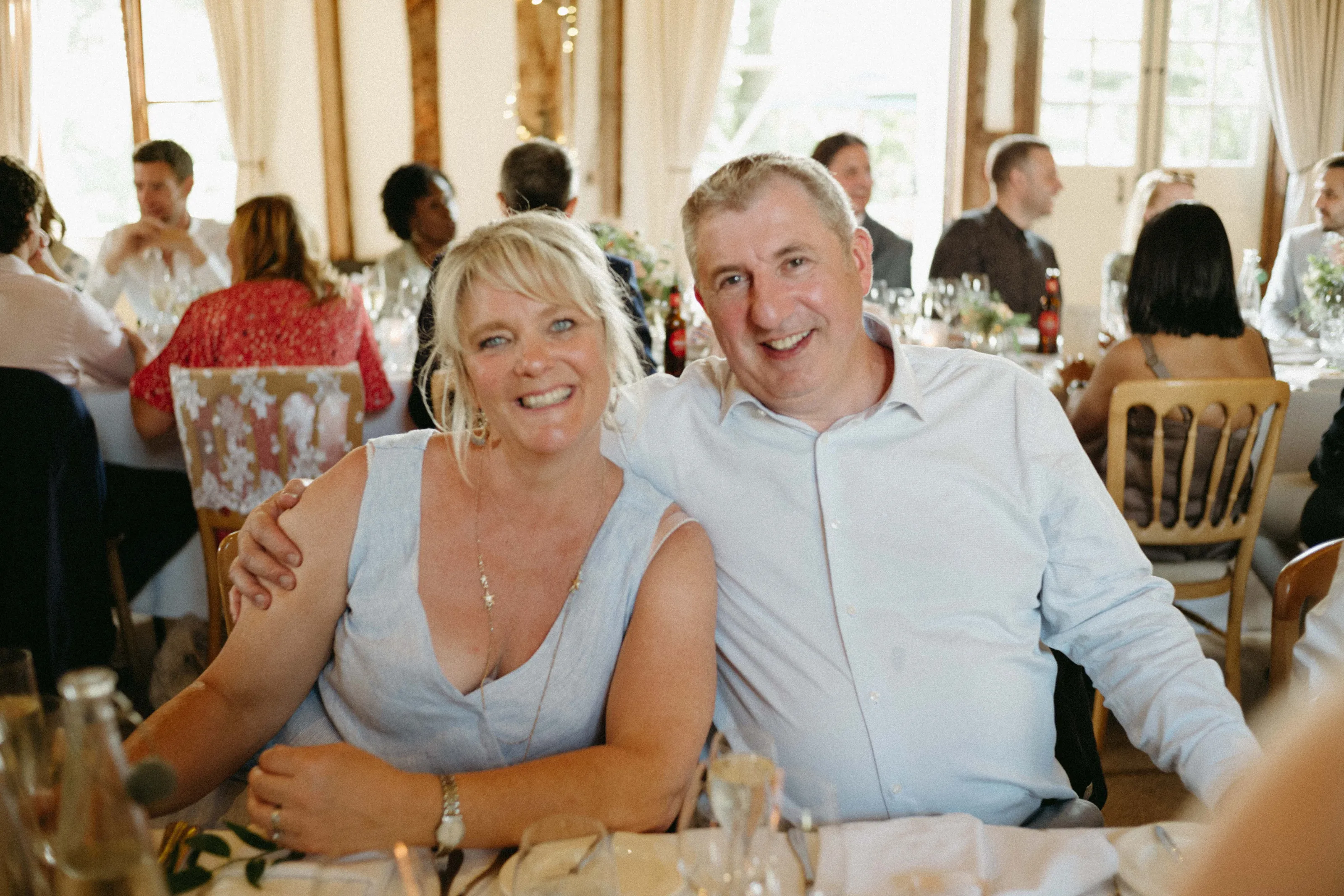 Smiling couple at a wedding reception, surrounded by guests.